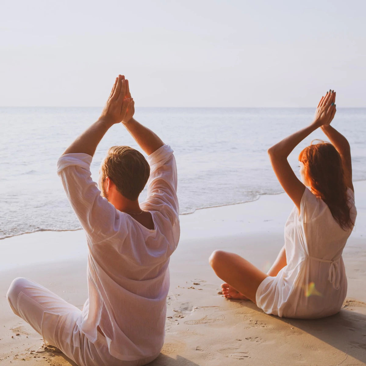 Man and lady practicing yoga on a beach with their hands above their heads in anjali mudra, whilst sitting cross legged in the easy pose or sukhasana