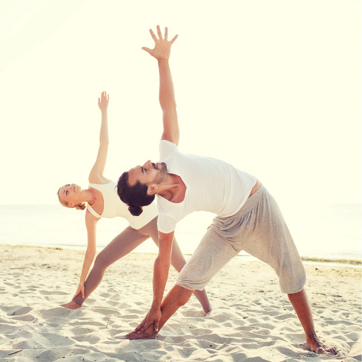 Man and lady on a beach practicing yoga, performing the yoga posture trikonasana, the triangle pose