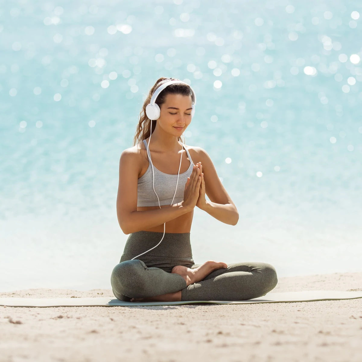Lady practicing yoga on a beach, wearing headphones performing the yoga posture ardha padmasana, the half lotus pose